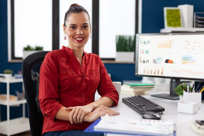 Smiling woman in office sitting at desk with computer screen illustrates blog "Accounts Receivable vs Control Account: What Is the Difference?"