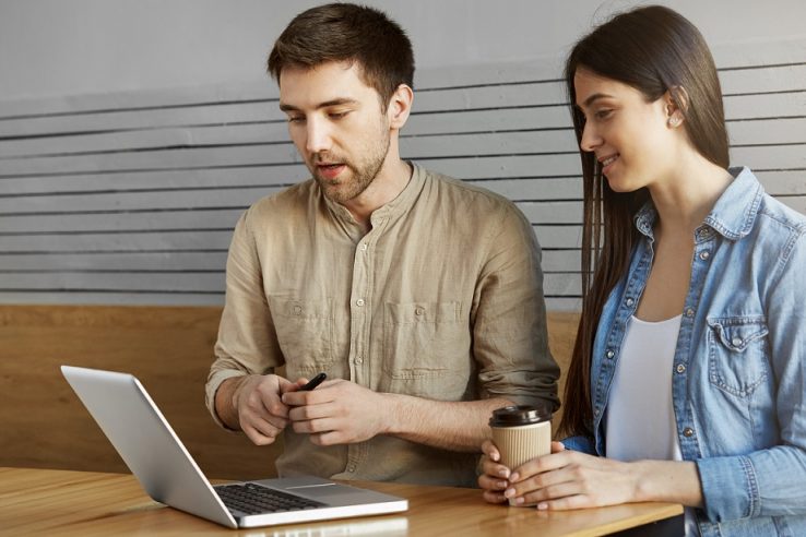 Two young perspective startup enthusiast sitting in cafe, drinking coffee talking about work and looking through project details on laptop computer. Relaxive and productive time Two casual people in office illustrate blog "Factoring vs Assignment of Receivables: What is the Difference?"