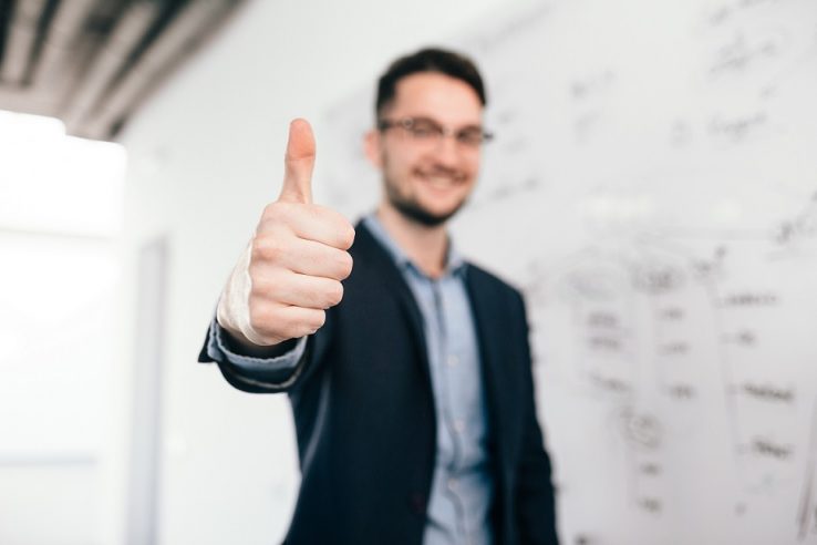 Young dark-haired man in glasses is standing near whiteboard in office. He wears blue shirt and dark jacket. Focus on front on his sign with hand Man giving thumbs up illustrates blog "What Does 'Cash Flow-Positive' Mean?"