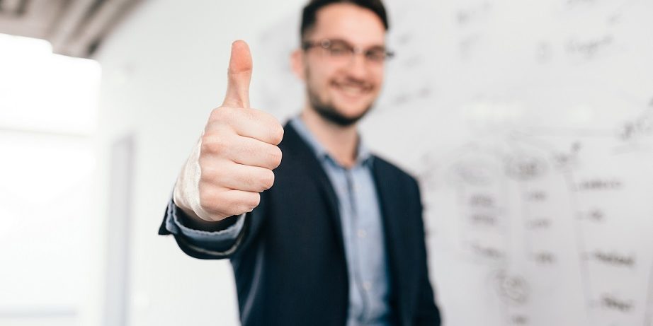 Young dark-haired man in glasses is standing near whiteboard in office. He wears blue shirt and dark jacket. Focus on front on his sign with hand Man giving thumbs up illustrates blog "What Does 'Cash Flow-Positive' Mean?"